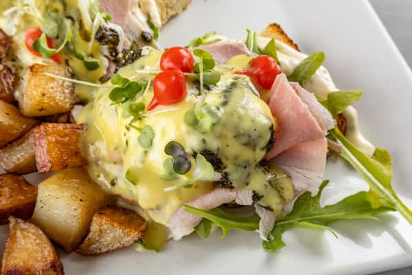 Close-up of eggs Benedict with thick-cut ham, hollandaise sauce, cherry tomatoes, and microgreens, served with a generous side of golden home fries.