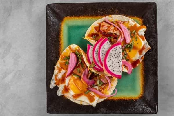 Close-up of vibrant huevos rancheros on a green and black ceramic plate, topped with fried eggs, salsa, bacon, pickled red onions, fresh cilantro, and slices of dragon fruit for a bold and flavorful Latin-inspired brunch dish.