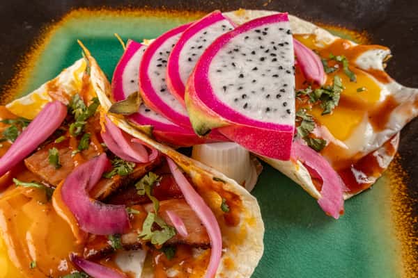 Bold huevos rancheros on a green ceramic plate, topped with fried eggs, salsa, bacon, pickled onions, fresh cilantro, and vibrant slices of dragon fruit for a colorful Latin-inspired brunch dish.