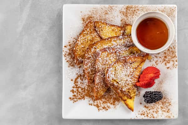 Golden French toast slices sprinkled with powdered sugar and cinnamon, served with a side of maple syrup, sliced strawberries, and a blackberry on a square white plate.