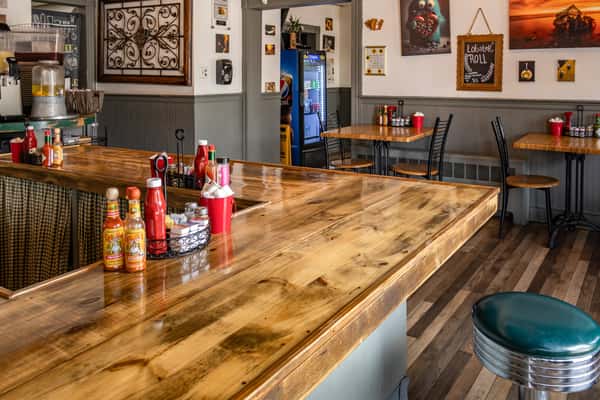 Cozy interior of Early Bird brunch restaurant featuring a polished wooden counter with classic diner stools, condiment caddies with hot sauce and ketchup, and wall art including a chalkboard sign advertising lobster rolls; small dining tables line the walls beneath framed artwork and gray wainscoting for a warm, casual dining atmosphere.