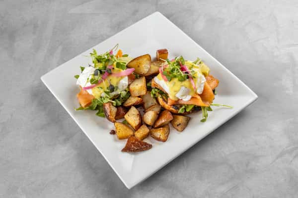Full plate of smoked salmon Benedict featuring arugula, poached eggs, and hollandaise over English muffins, served with crispy roasted breakfast potatoes on a square white plate.