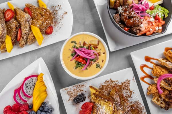 Overhead view of a diverse brunch sampler including French toast with fruit, a skillet hash with eggs and vegetables, cheesy queso dip topped with pickled onions, and a colorful fruit platter for a bold and indulgent brunch feast.