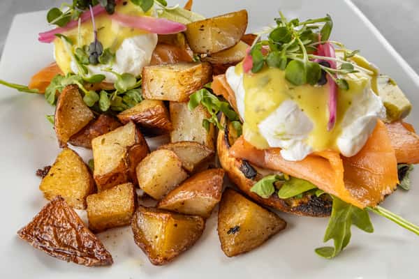 Smoked salmon eggs Benedict with poached eggs and hollandaise sauce, garnished with microgreens and pickled onions, served over toasted English muffins with a side of golden-brown home-fried potatoes.