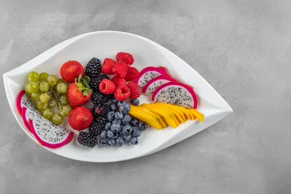 Tropical fruit platter served on a white leaf-shaped dish, featuring vibrant slices of dragon fruit, ripe mango, strawberries, green grapes, blackberries, blueberries, and raspberries for a colorful and refreshing brunch option.