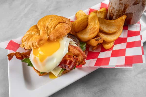 Flaky croissant sandwich filled with bacon, egg, and cheese, served with seasoned curly fries and a chocolate shake on red-and-white checkered paper for a classic American brunch combo.