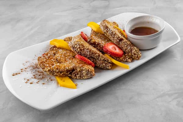 Crunchy, nut-crusted French toast slices dusted with powdered sugar and cinnamon, layered with sliced strawberries and mango, and served with a side of maple syrup on a rectangular white plate.