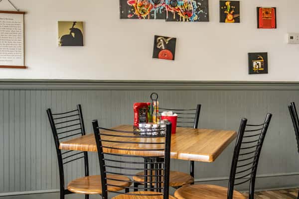 Casual dining area at Early Bird restaurant featuring a wooden table set for four, black metal chairs, a condiment caddy, and eclectic modern artwork against sage green wainscoting.