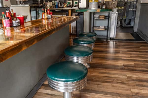 Retro-style diner counter with polished wood surface and teal vinyl swivel stools, set against warm wood flooring and a classic setup of condiments, napkin holders, and a drink station for a nostalgic, casual brunch atmosphere.