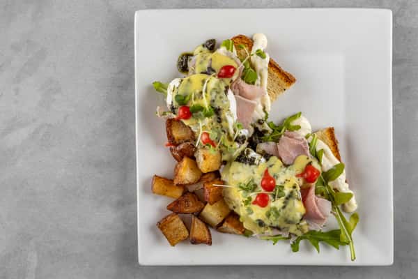 Toasted bread topped with poached eggs, sliced ham, fresh greens, and creamy hollandaise sauce, served alongside crispy home-fried potatoes for a hearty brunch plate.