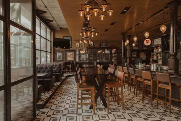Spacious Irish pub bar with high-top tables, leather bar stools, patterned tile flooring, and hanging pendant lights, designed for casual dining and drinks.