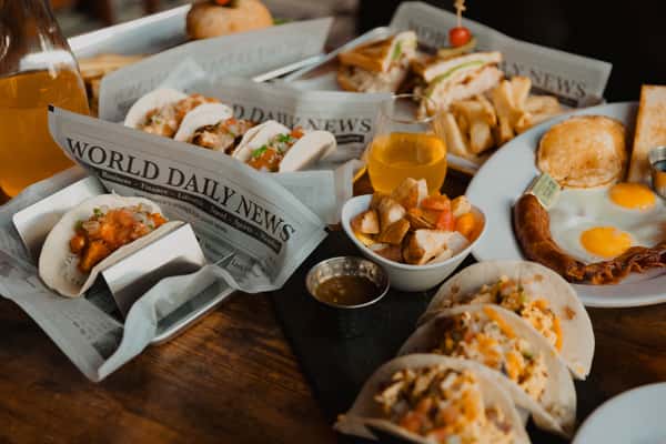 Irish pub brunch spread featuring street-style tacos, toasted sandwiches with fries, breakfast plates with eggs and sausage, and mimosas served on a wooden table.
