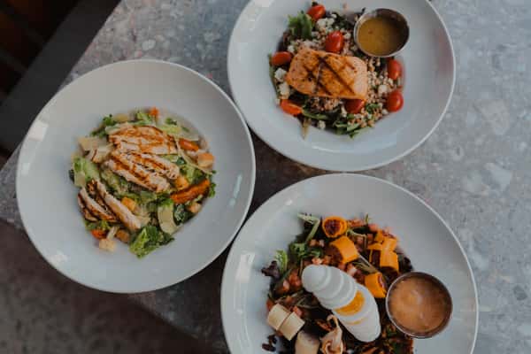 Three fresh entrée salads topped with grilled chicken and salmon, mixed greens, roasted vegetables, and house dressings at an Irish pub restaurant.