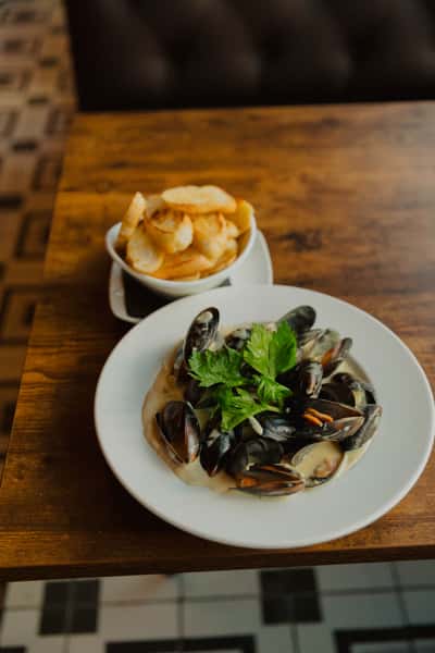 Steamed mussels in a creamy broth garnished with fresh herbs, served with house-made chips on a wooden pub table.