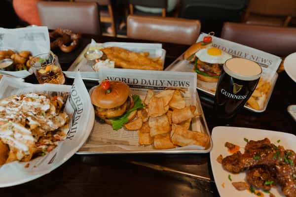 Large Irish pub food spread featuring burgers, fish and chips, wings, nachos, and a pint of Guinness, arranged family-style on a dining table.