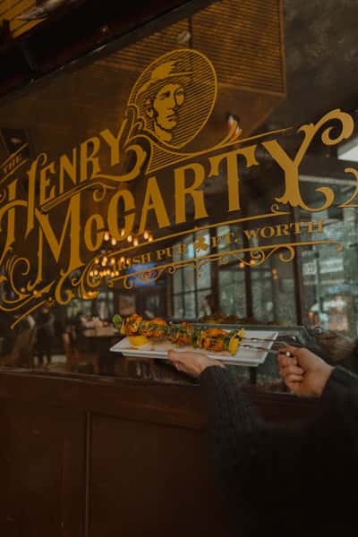 Hands serving grilled skewers in front of The Henry McCarty Irish Pub glass signage, highlighting signature pub dishes and an inviting Fort Worth bar setting.