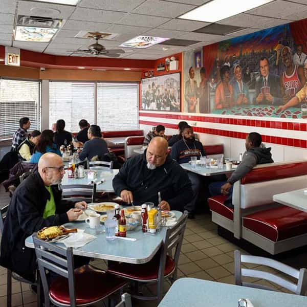 Busy retro-style diner with red booths and multiple groups of people eating and talking at tables, with a large mural on the wall.