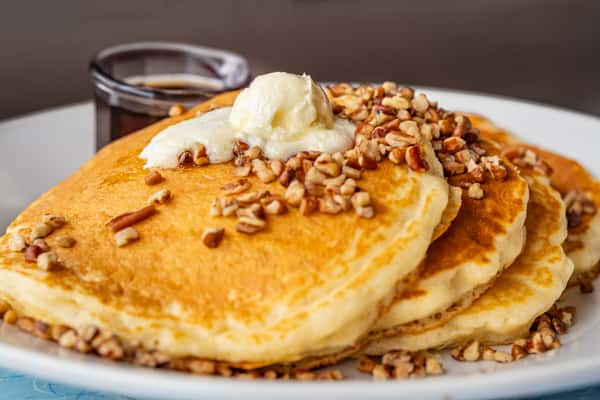 Stack of pancakes topped with butter and chopped pecans, served with a side of syrup.