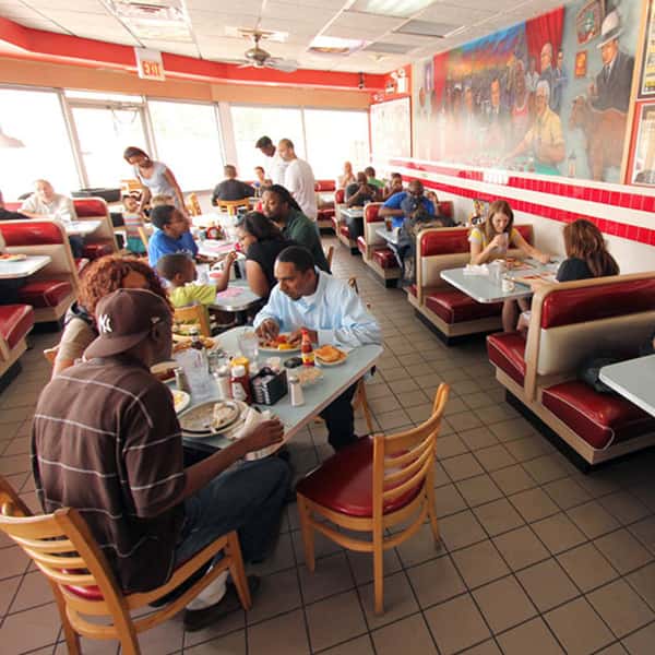 Busy diner scene with red booths and several groups of people eating and talking at tables near large windows and a wall mural.