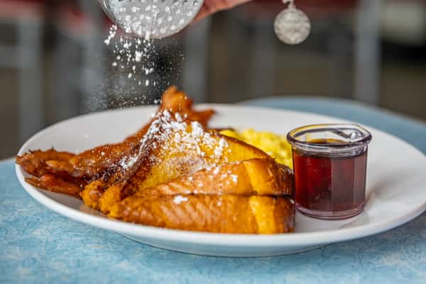 French toast topped with powdered sugar, served with bacon, scrambled eggs, and a side of syrup.