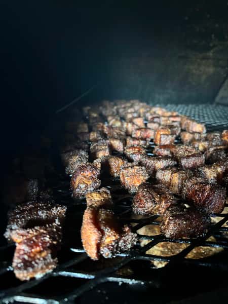 The image features small pieces of meat, likely burnt ends or cubed brisket, cooking on a grill with a smoky haze in the background.