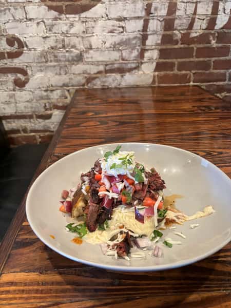 A plate of finely shredded beef topped with fresh tomato, onion, cilantro, and shredded cheese, sitting on a bed of cabbage. The background shows a brick wall.