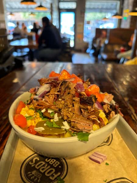 A close-up of a bowl filled with pulled pork, tomatoes, pickles, onions, cilantro, and other fresh ingredients, with a blurred background of a restaurant.