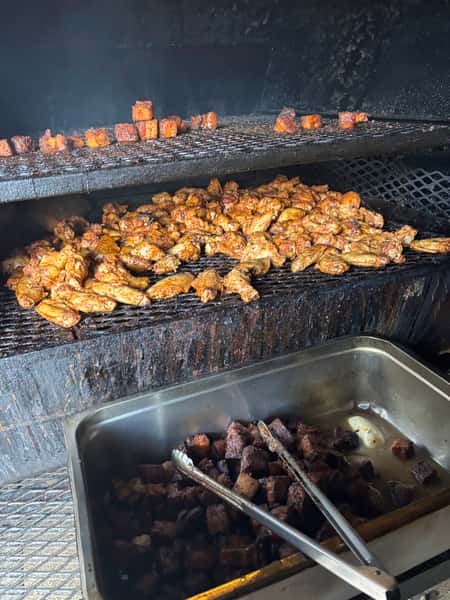 The image shows a smoker with barbecue chicken wings grilling on the bottom rack and small pieces of meat on the top rack. A tray below holds more cooked pieces.