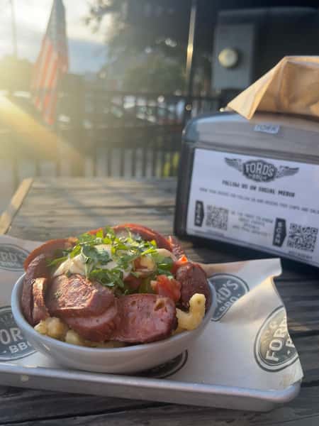 A bowl of sausage, macaroni, and cheese topped with fresh cilantro and tomatoes, served outdoors with a "Ford's BBQ" takeout box in the background.