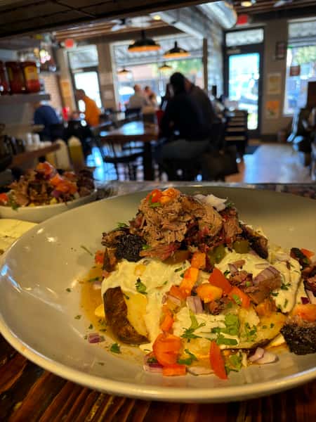 A loaded baked potato topped with pulled pork, cheese, sour cream, diced onions, tomatoes, jalapeños, and chopped herbs, served in a white bowl inside a cozy barbecue restaurant.