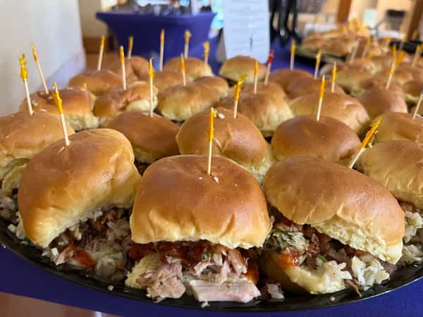 A platter of pulled pork sandwiches, each topped with a toothpick, filled with meat, slaw, and barbecue sauce in soft buns.