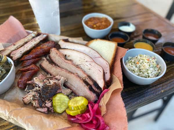 A plate of smoked meats including pulled pork, sausage, brisket, and turkey, accompanied by pickles, coleslaw, cornbread, and various sauces.