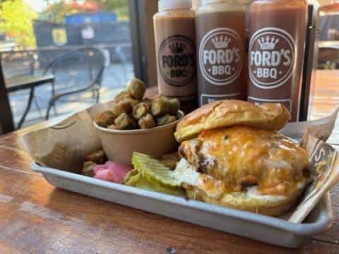 A BBQ sandwich with melted cheese, pickles, and a side of fried okra served with bottles of Ford's BBQ sauce.