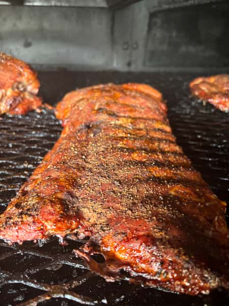 The image shows racks of ribs cooking on a grill, with a well-seasoned crust on the meat and a smoky atmosphere in the background.