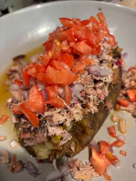 A loaded baked potato topped with pulled pork, diced tomatoes, and onions, drizzled with a savory sauce.