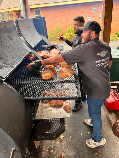 Two men tending to a large smoker with various cuts of meat cooking on it.
