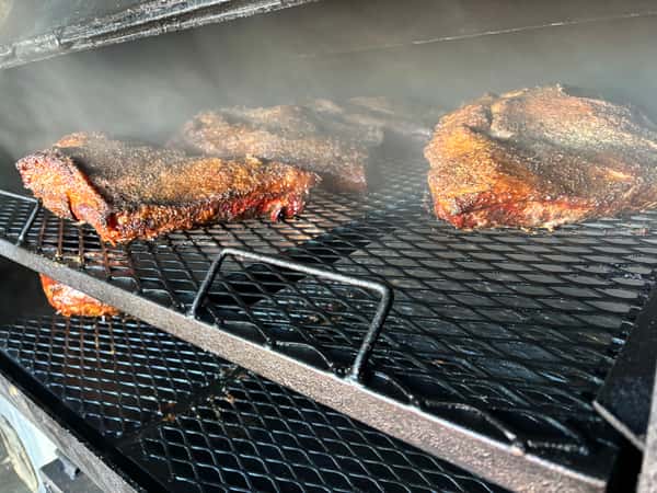 The image shows a close-up of several pieces of smoked meat, likely brisket, resting on a grill. The meat is coated with a dark, flavorful rub, and the smoke rising from the grill adds to the mouth-watering atmosphere of the scene.