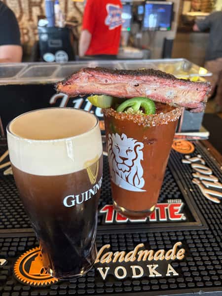 A pint of Guinness beer next to a spicy drink garnished with a rib, lime slices, and a salted rim, sitting on a bar mat. The background shows people and a bar counter.