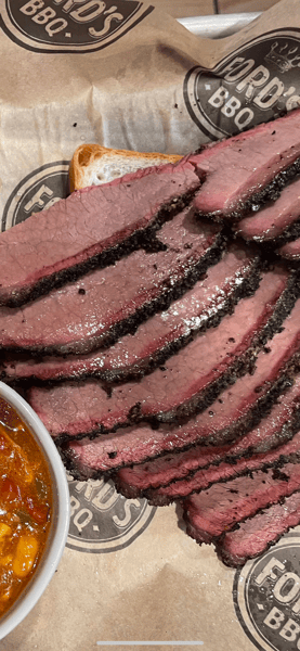 A close-up of tender, sliced brisket paired with a side of baked beans and served on butcher paper with a slice of bread.