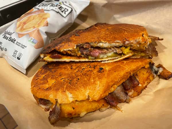 A delicious grilled sandwich filled with bacon, melted cheese, and slices of meat, with a side of potato chips.