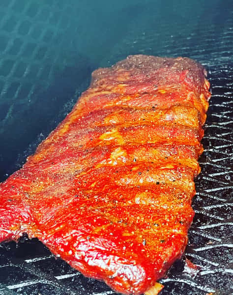 A close-up of a rack of ribs on a smoker, coated in a red BBQ glaze with visible grill marks and smoky texture.