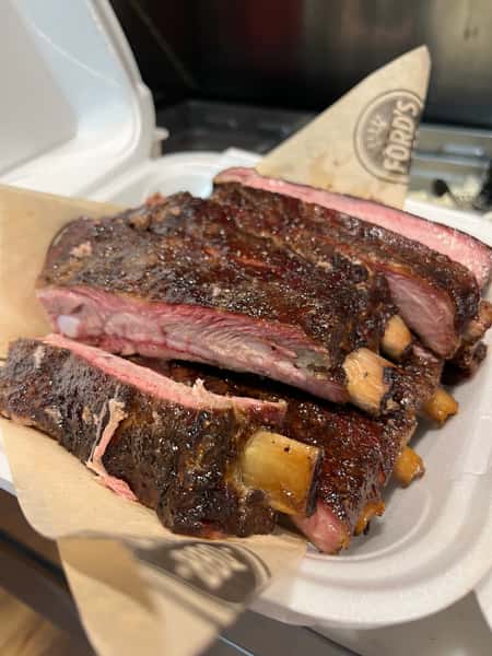 A close-up of tender, juicy ribs with a perfect char on the edges, served in a takeout container.