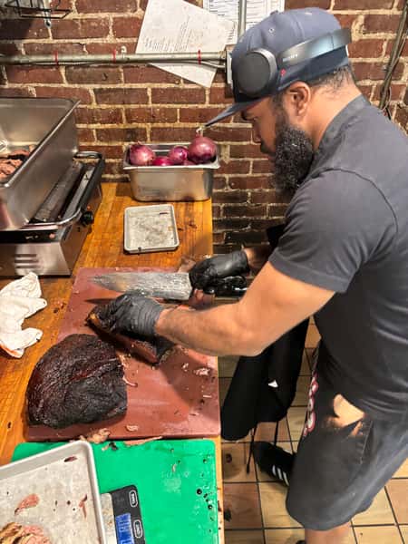 A man wearing gloves and headphones slices smoked brisket on a cutting board in a barbecue kitchen with onions and equipment nearby.