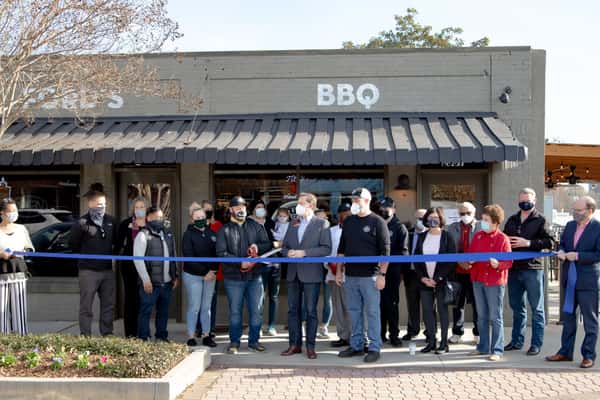 The image shows a ribbon-cutting ceremony in front of a BBQ restaurant. A group of people, including staff and officials, are gathered outside the restaurant with a large blue ribbon, marking the grand opening. Several individuals are wearing masks, and the restaurant's name is visible on the sign.