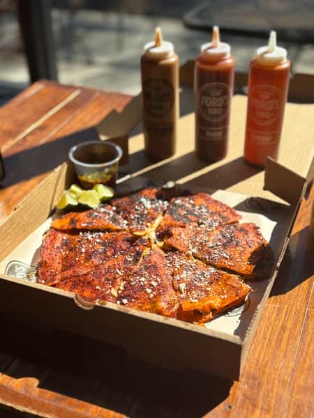 A large serving of crispy, smoky, seasoned quesadilla slices topped with cheese and served with pickles and barbecue sauce on the side.