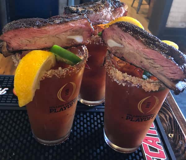 Three Bloody Mary drinks garnished with a slice of smoked brisket, orange wedge, and green jalapeño.