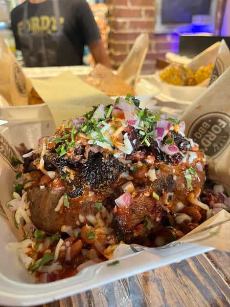 A baked potato topped with rice, beans, pulled pork, cheese, diced onions, cilantro, and BBQ sauce, served in a takeout container.