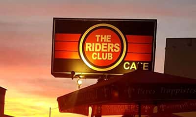 Illuminated sign for The Riders Club Café glowing at sunset, with bold red and orange stripes and a vintage aesthetic, positioned above a patio umbrella.