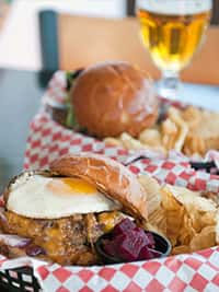 Close-up of a gourmet cheeseburger topped with a fried egg, served in a red checkered basket with kettle chips and pickled beets, with a second burger and a glass of beer in the background at a casual American diner.