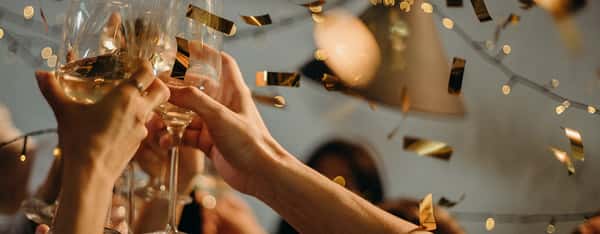 Group of hands toasting with wine glasses under warm festive lights and gold confetti, capturing a lively celebration or special event ambiance.